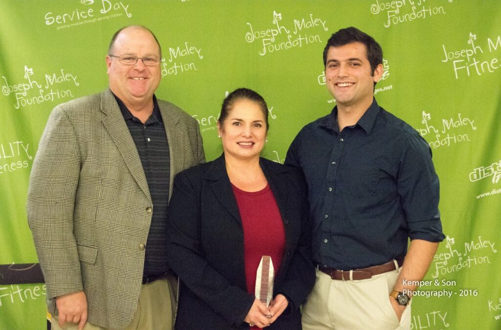 Ivette Bruns standing between her husband on the left and her son on the right in front of the green JMF step and repeat banner