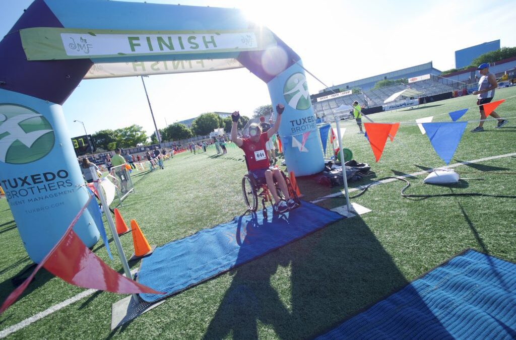 Man in wheelchair crossing the finish line at the JMF 5k walk run roll raising his hands in victory