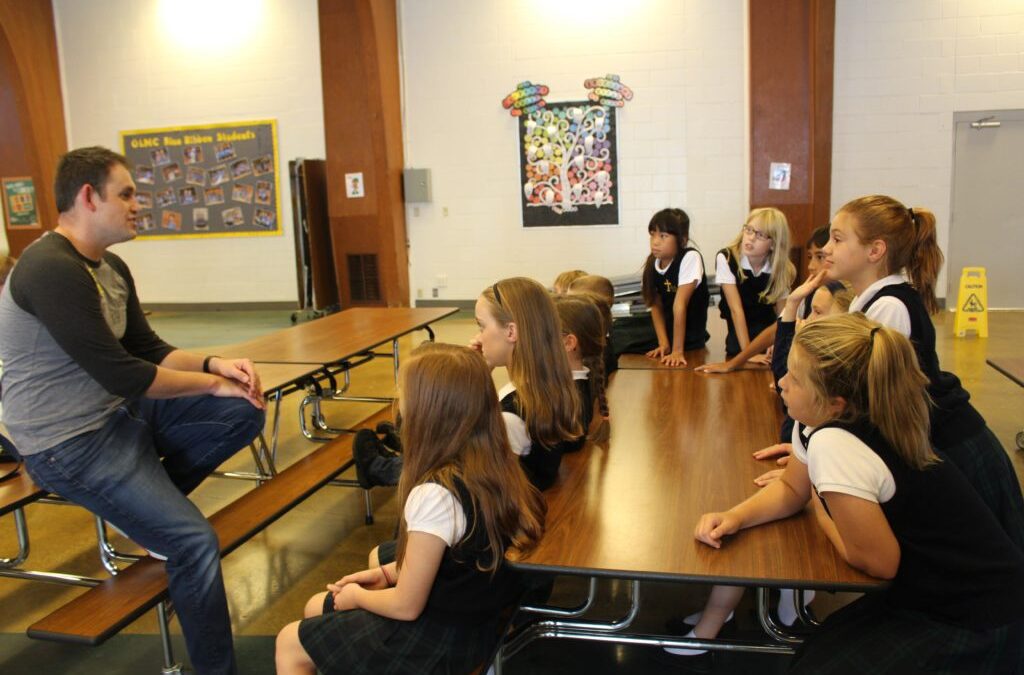 Adult male speaking to a group of school girls dressed in school uniforms and sitting at a long table