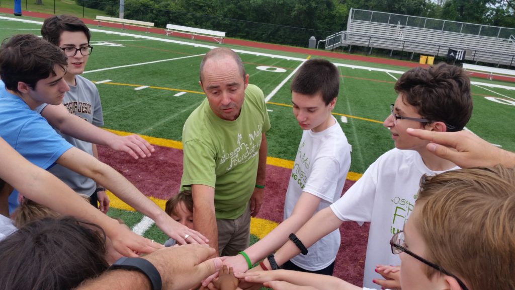 Adult male wearing green JMF shirt leading a huddle of teen-aged boys on a football field