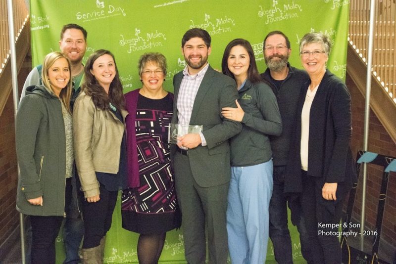 9 family-friends smiling in front of green JMF step and repeat banner to honor man in middle holding an award