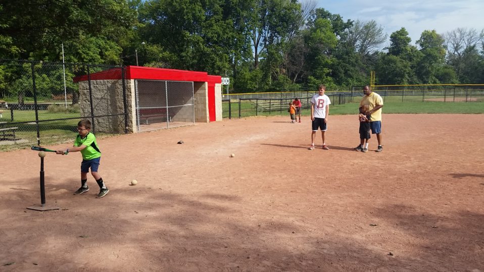kid hitting t-Ball on dirt field with some spectators in the back