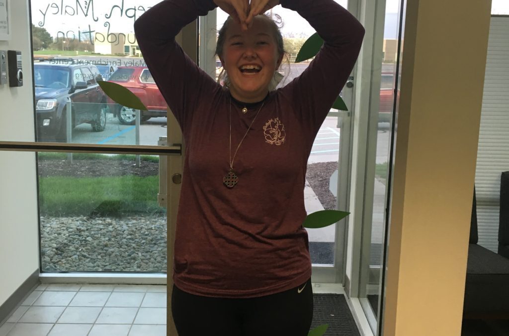 Emily Schommer posing in front of glass doors at JMF and making a heart shape with her arms