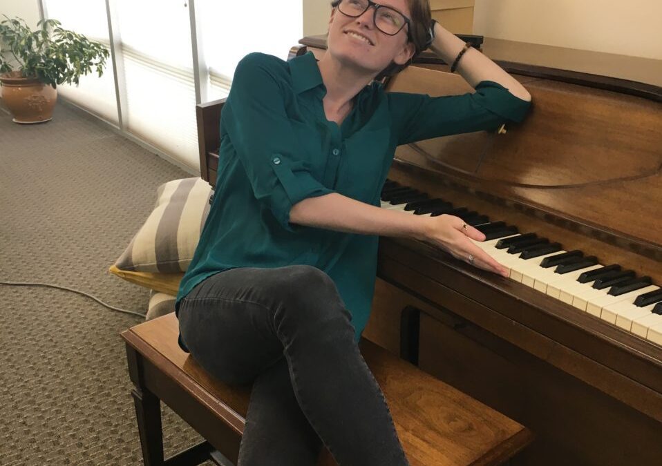 Molly Dennie wearing teal button-down shirt and jeans and posing playfully on a piano bench