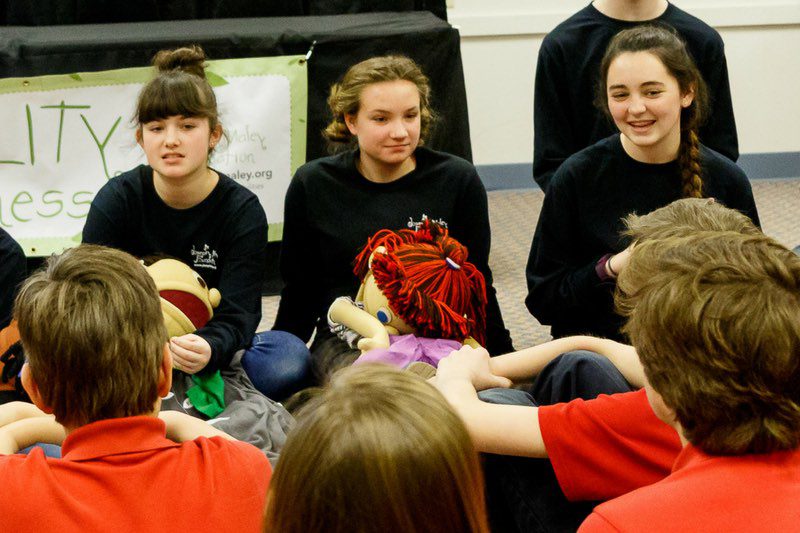 girls sitting on a classroom floor with puppets on their laps and kids on the opposite side listening