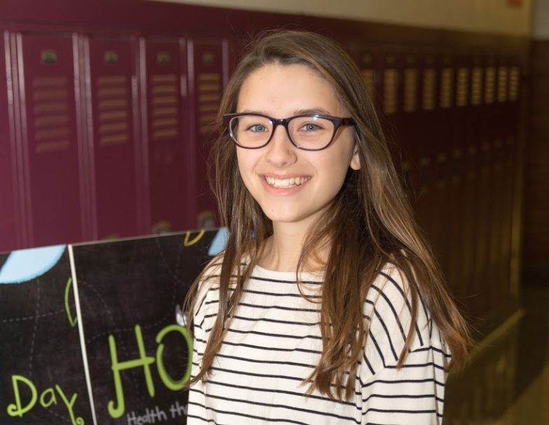 Aubrey Weist wearing striped shirt and glasses while smiling for a picture in front of red lockers and JMF signs peeking through