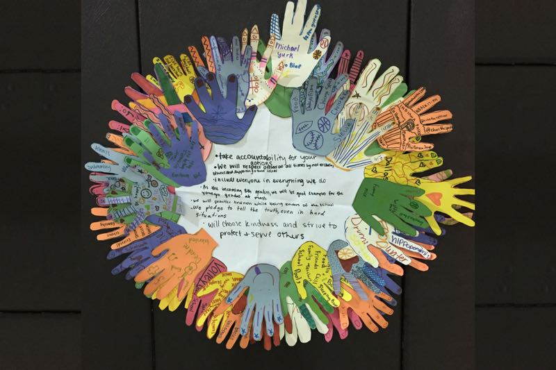 cut-out paper hands of varying colors placed in a circle on a black background to form a wreath with message in the middle