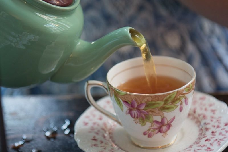 Image of a green teapot pouring into a vintage white teacup with pink flowers on it.