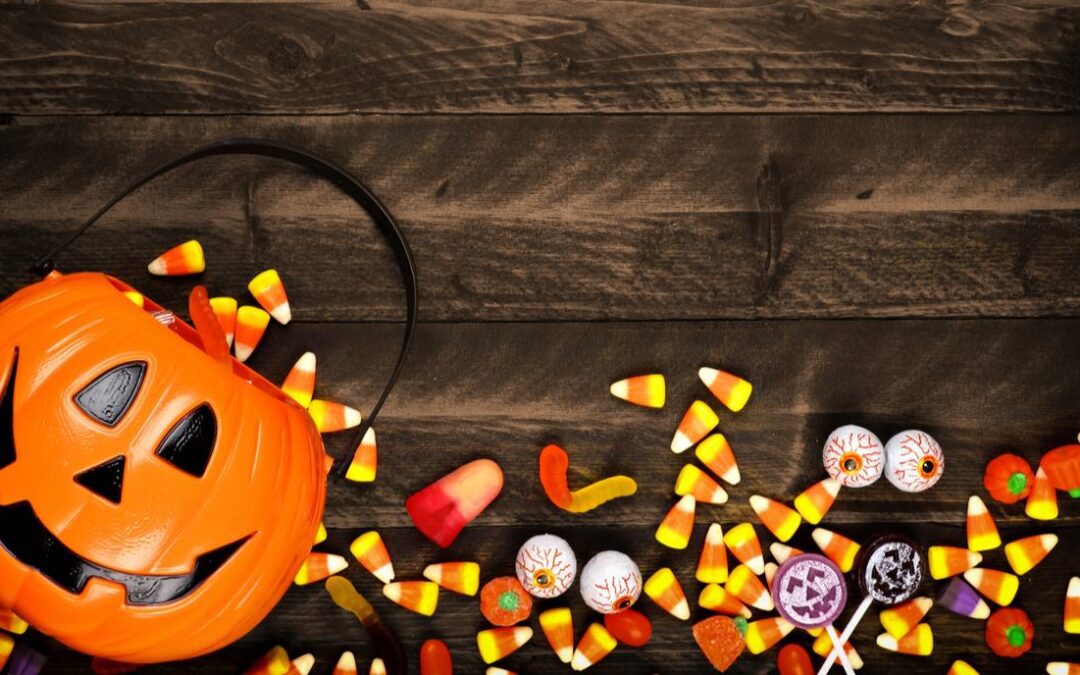 Halloween candy spread across the bottom of a wooden table and a plastic pumpkin container on the left